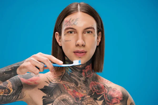 Hombre joven y tatuado sosteniendo cepillo de dientes aislado en azul - foto de stock