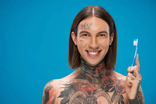 Uomo allegro e tatuato che tiene lo spazzolino da denti mentre sorride isolato sul blu — Foto stock