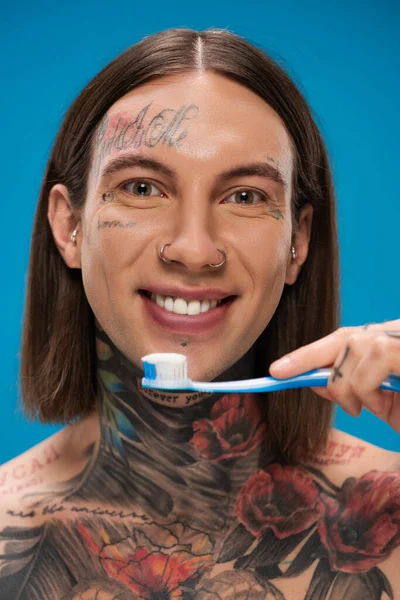 Positive and tattooed man holding toothbrush while smiling isolated on blue — Stock Photo