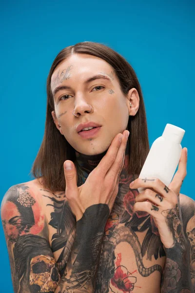 Homme tatoué tenant bouteille avec un produit cosmétique et toucher la peau lisse isolé sur bleu — Photo de stock