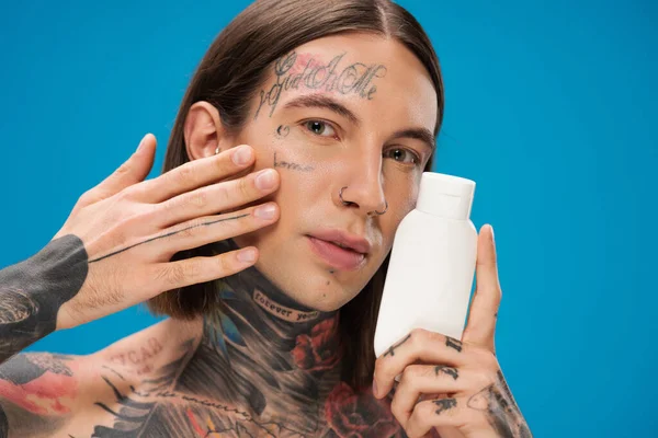 Jeune homme percé tenant bouteille avec un produit cosmétique et toucher la peau lisse isolé sur bleu — Photo de stock