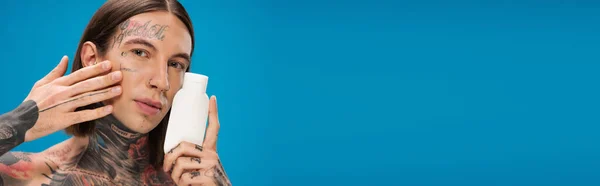 Пирсинг и татуировка мужчина держит бутылку с косметическим продуктом и прикосновение гладкой кожи изолированы на синий, баннер — стоковое фото