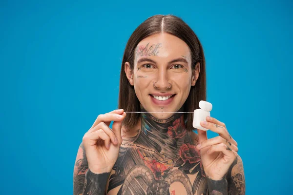 Cheerful and tattooed man holding dental floss while looking at camera isolated on blue — Stock Photo