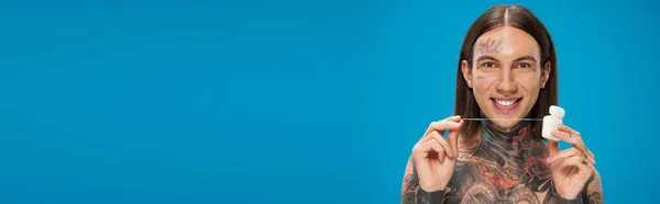 Cheerful and tattooed man holding dental floss isolated on blue, banner — Stock Photo