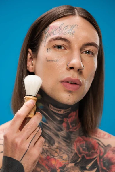 Young and tattooed man holding shaving brush near face isolated on blue — Stock Photo