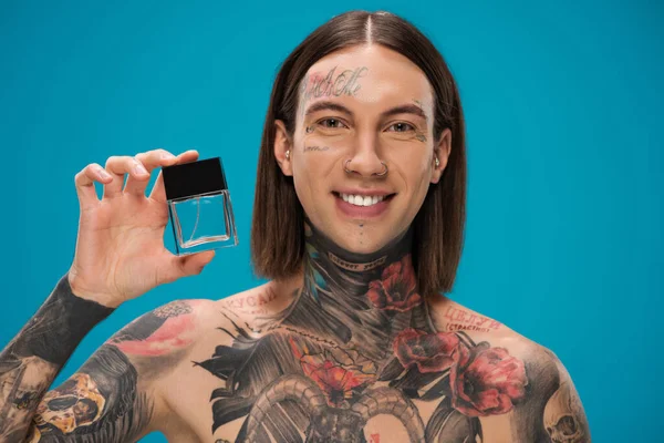 Hombre feliz y tatuado sosteniendo botella con perfume aislado en azul - foto de stock