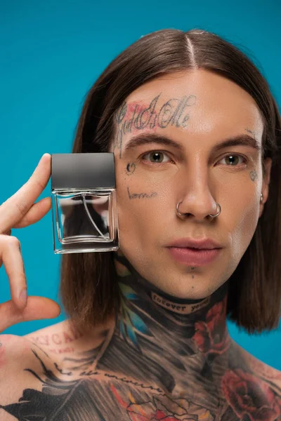 Modelo joven y tatuado sosteniendo botella con perfume aislado en azul - foto de stock