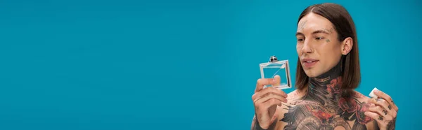 Joven y tatuado hombre mirando botella con perfume aislado en azul, bandera — Stock Photo