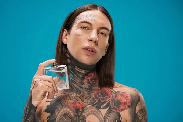 Hombre joven y tatuado sosteniendo botella y rociando perfume aislado en azul — Stock Photo