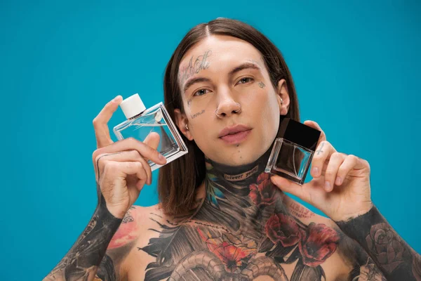Hombre joven y tatuado sosteniendo botellas con perfume aislado en azul — Stock Photo