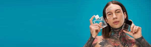 Hombre joven y tatuado sosteniendo botellas con perfume aislado en azul, pancarta — Stock Photo