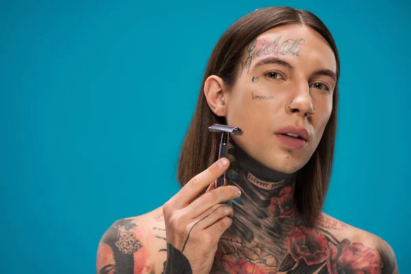Татуированный молодой человек с пирсингом держа безопасную бритву и бритье лицо изолированы на синий — стоковое фото