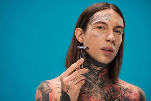 Tattooed young man with piercing holding safety razor and shaving face isolated on blue — Stock Photo