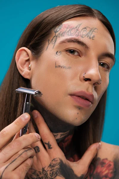 Primo piano di giovane uomo con tatuaggi in possesso di rasoio di sicurezza e faccia da barba isolato su blu — Foto stock