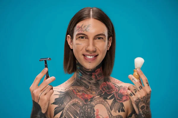 Happy young man with tattoos holding safety razor and vintage shaving brush isolated on blue — Stock Photo