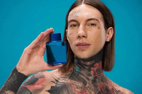 Jovem com tatuagens segurando garrafa com após barbear isolado em azul — Fotografia de Stock