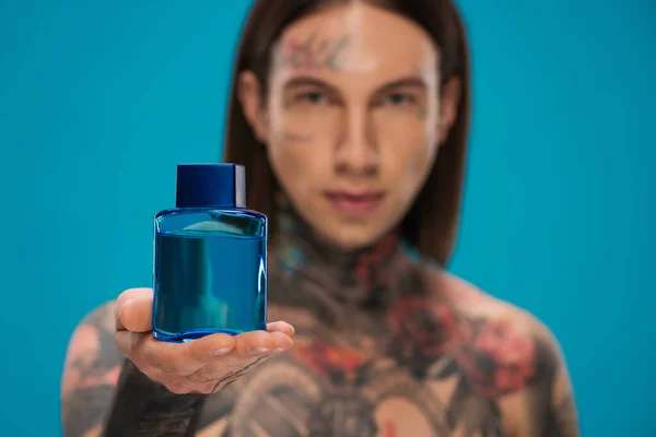 Jeune homme flou avec tatouages tenant bouteille avec après-rasage isolé sur bleu — Photo de stock