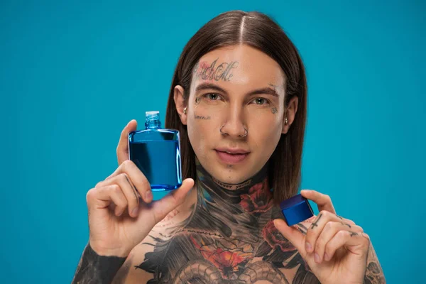 Pierced young man with tattoos holding bottle with after shave isolated on blue — Stock Photo