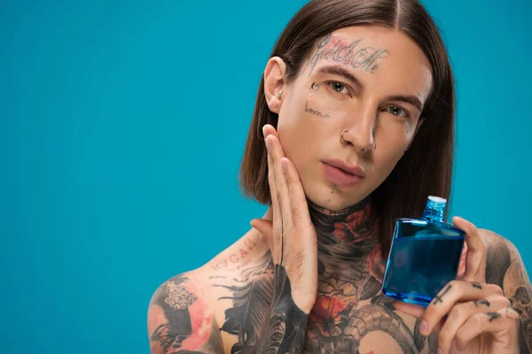 Jeune homme avec des tatouages tenant bouteille tout en appliquant après rasage isolé sur bleu — Photo de stock