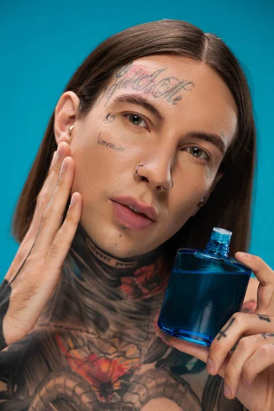 Joven con tatuajes aplicando después de afeitarse en la mejilla aislado en azul - foto de stock