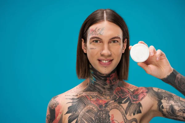 Feliz joven con tatuajes sosteniendo contenedor con crema facial aislado en azul - foto de stock