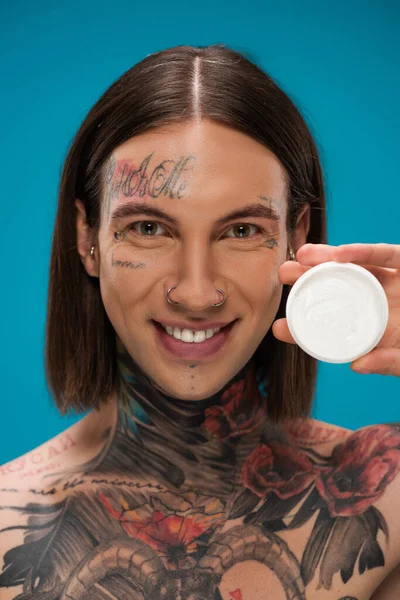 Homem jovem positivo segurando recipiente com creme cosmético isolado em azul — Fotografia de Stock