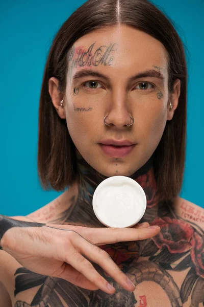 Jovem perfurado com tatuagens segurando recipiente com creme cosmético isolado em azul — Fotografia de Stock