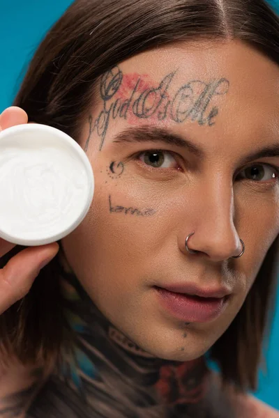 Close-up de jovem tatuado segurando recipiente com creme cosmético isolado em azul — Fotografia de Stock