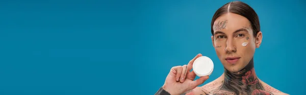 Tattooed young man with cream on cheek holding container isolated on blue, banner — Stock Photo