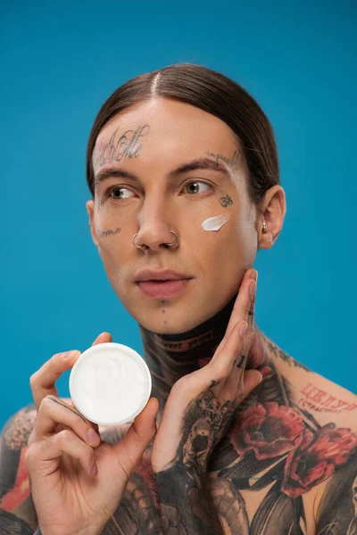 Tattooed young man with face cream on cheek holding container isolated on blue — Stock Photo
