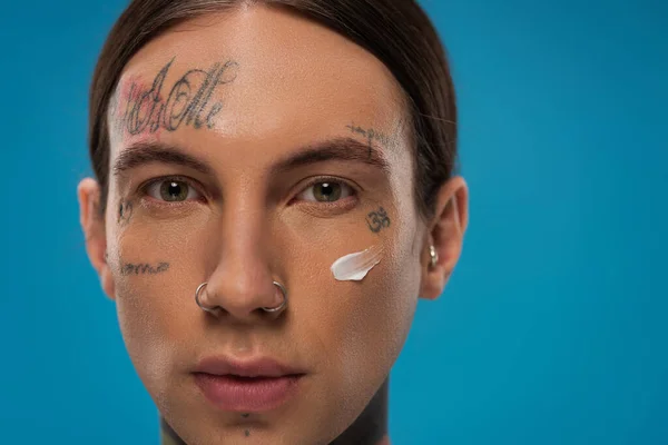 Primo piano del giovane tatuato con crema viso sulla guancia guardando la fotocamera isolata sul blu — Foto stock