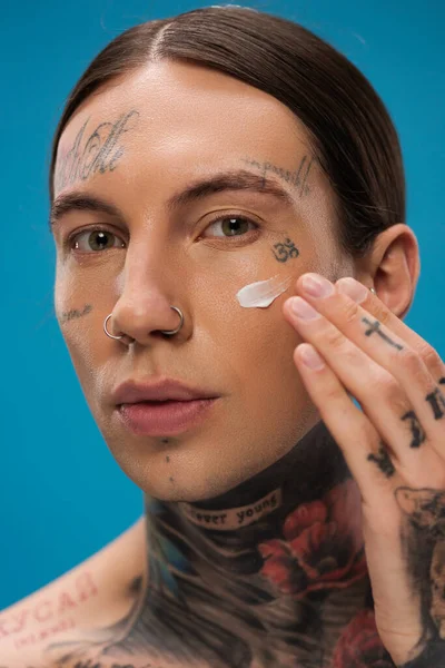 Tattooed young man applying face cream on cheek and looking at camera isolated on blue — Stock Photo