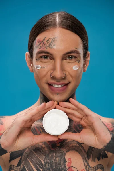 Happy young man with tattoos and cream on cheeks holding container isolated on blue — Stock Photo