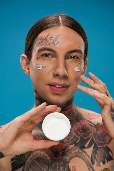Jovem tatuado com creme cosmético nas bochechas segurando recipiente isolado em azul — Fotografia de Stock