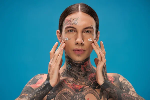 Hombre joven tatuado aplicando crema en las mejillas mientras mira la cámara aislada en azul - foto de stock