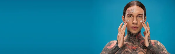Tatuato giovane uomo applicando crema sulle guance, mentre guardando la fotocamera isolata su blu, banner — Foto stock