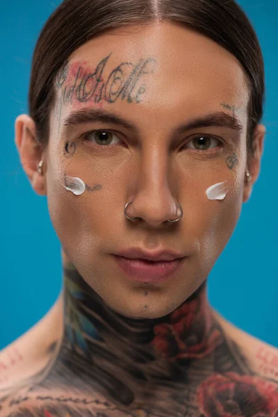 Hombre joven tatuado con crema facial en las mejillas mirando la cámara aislada en azul - foto de stock