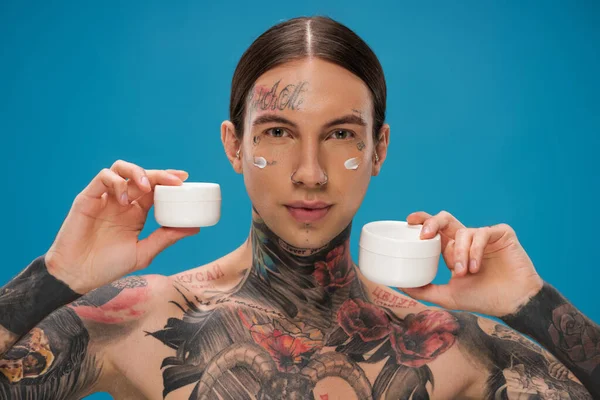 Tätowierter junger Mann mit verschiedenen Behältern mit Kosmetikcreme auf blauem Grund — Stockfoto