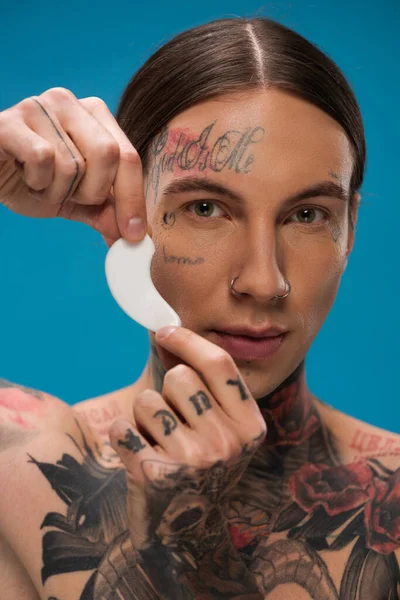 Joven tatuado sosteniendo parche en los ojos y mirando a la cámara aislada en azul - foto de stock