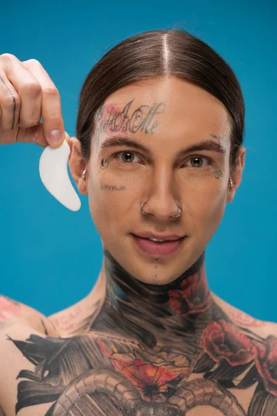 Tattooed young man holding eye patch and smiling while looking at camera isolated on blue — Stock Photo