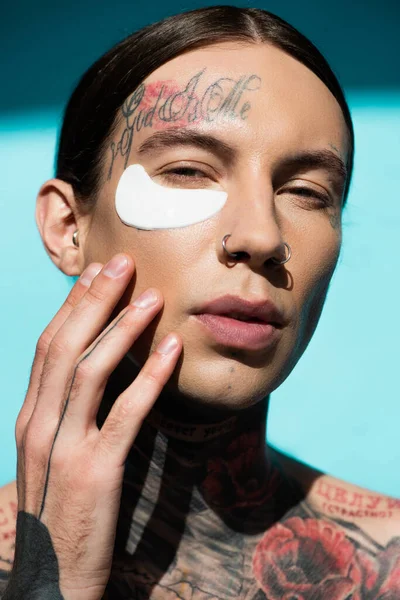 Joven tatuado aplicando parche en los ojos y mirando a la cámara en turquesa - foto de stock