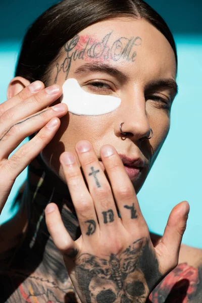 Tattooed young man applying eye patch with hands and looking at camera on turquoise — Stock Photo