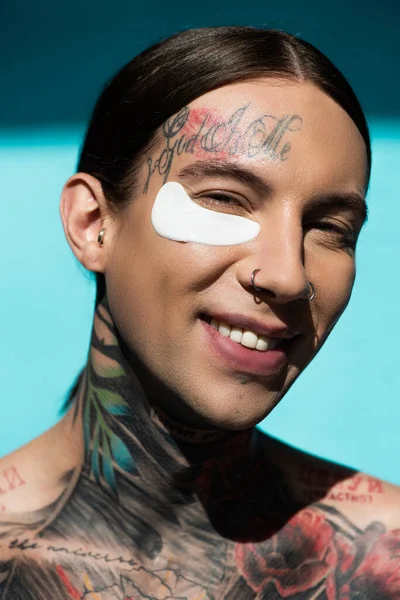 Tattooed young man with eye patch smiling while looking at camera on turquoise — Stock Photo