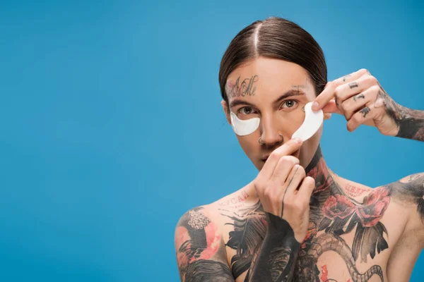 Joven con tatuajes aplicando parches en los ojos y mirando a la cámara aislada en azul - foto de stock