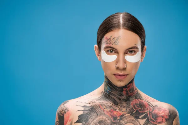 Hombre joven sin camisa y tatuado con parches en los ojos mirando a la cámara aislada en azul - foto de stock