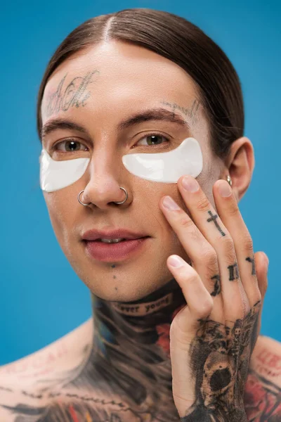 Gros plan de jeune homme souriant avec des tatouages et des patchs pour les yeux en regardant la caméra isolée sur bleu — Photo de stock