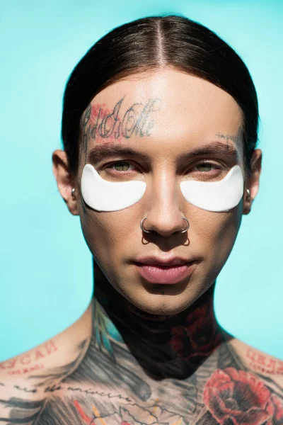 Gros plan de jeune homme tatoué avec des bandeaux oculaires regardant la caméra isolée sur turquoise — Photo de stock