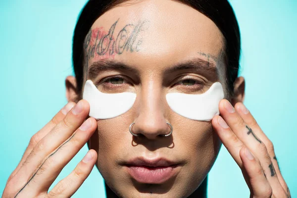 Close up de tatuado jovem homem ajustando manchas nos olhos e olhando para a câmera isolada em turquesa — Fotografia de Stock