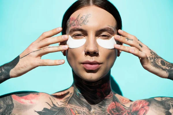 Jeune homme torse nu et tatoué avec des patchs pour les yeux regardant la caméra sur turquoise — Photo de stock