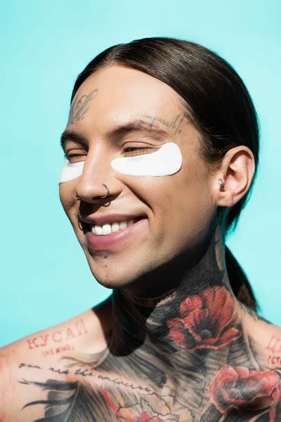 Joven feliz y tatuado con parches en los ojos sonriendo aislado en turquesa - foto de stock